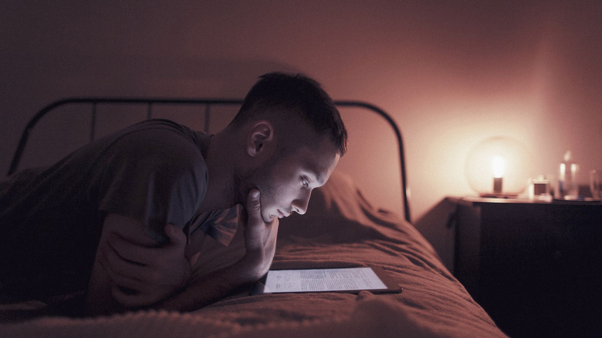 Photo of young man on his bed, reading a tablet.